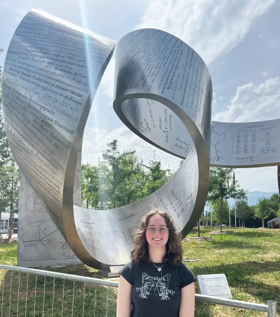 Photo: Taylor in front of "Wandering the Immeasurable," a sculpture designed by Gayle Hermick that welcomes CERN visitors. From the Mesopotamians' cuneiform script to the mathematical formalism behind the discovery of the Higgs boson, the sculpture narrates the story of how knowledge is passed through the generations and illustrates the aesthetic nature of the mathematics behind physics. (Description Credit: ATLAS experiment)

